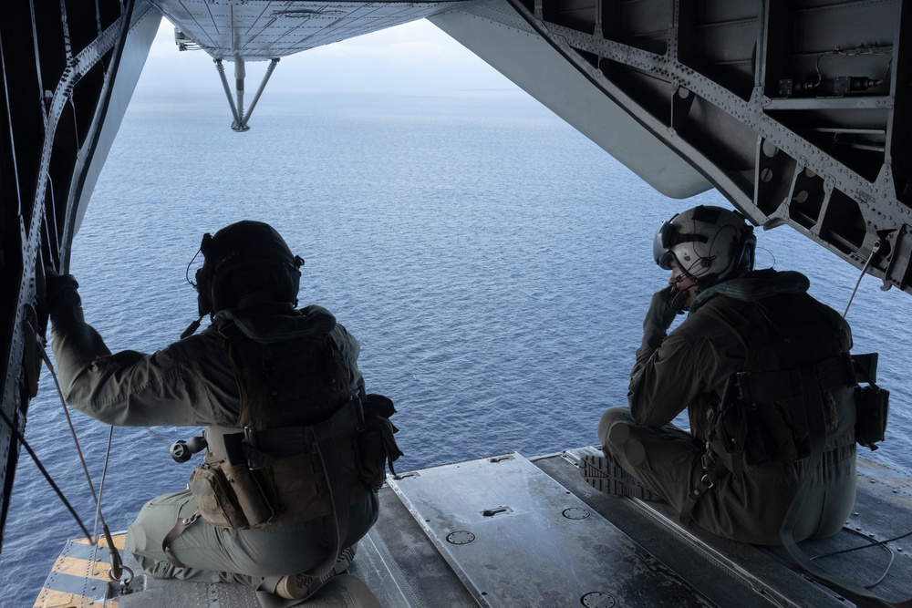Two military personnel in full tactical gear are seated on the open rear ramp of a military aircraft, looking out over a large body of open water. Both individuals are wearing flight helmets, body armor, and tactical vests. The image is taken from inside the aircraft looking outward toward the ocean horizon.