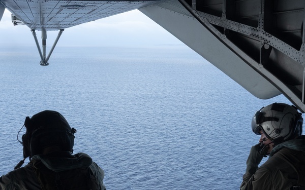 Two military personnel in full tactical gear are seated on the open rear ramp of a military aircraft, looking out over a large body of open water. Both individuals are wearing flight helmets, body armor, and tactical vests. The image is taken from inside the aircraft looking outward toward the ocean horizon.