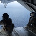 Two military personnel in full tactical gear are seated on the open rear ramp of a military aircraft, looking out over a large body of open water. Both individuals are wearing flight helmets, body armor, and tactical vests. The image is taken from inside the aircraft looking outward toward the ocean horizon.