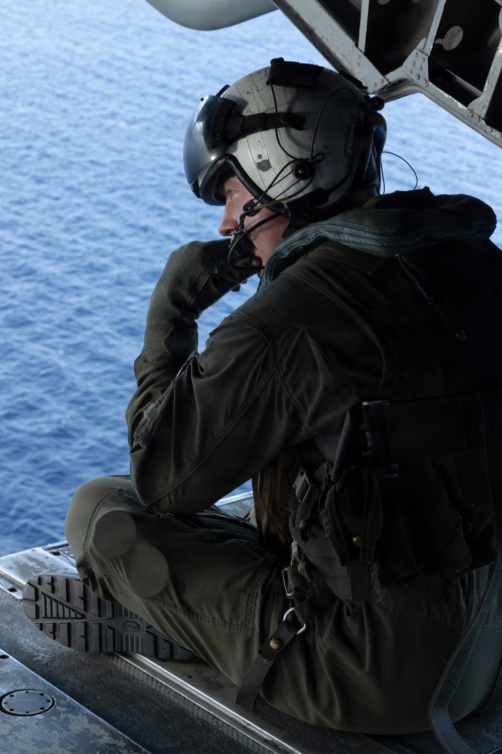 A military aircrew member is seated on the open ramp or door of an aircraft in flight over open ocean. The individual is wearing full flight gear including a helmet with visor, flight suit, and communications equipment, and appears to be scanning or observing the water below. The blue ocean is clearly visible in the background, suggesting the aircraft is at a moderate altitude.