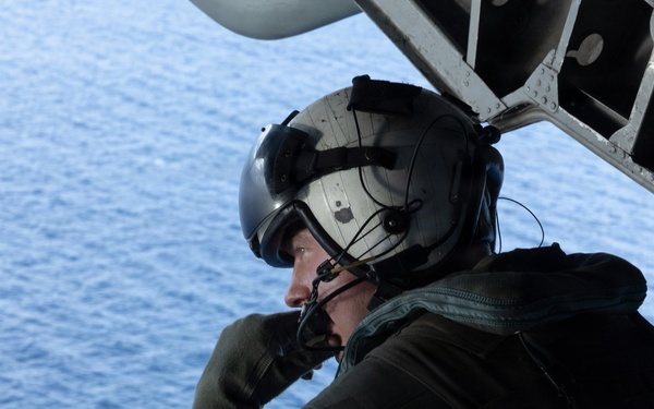 A military aircrew member is seated on the open ramp or door of an aircraft in flight over open ocean. The individual is wearing full flight gear including a helmet with visor, flight suit, and communications equipment, and appears to be scanning or observing the water below. The blue ocean is clearly visible in the background, suggesting the aircraft is at a moderate altitude.