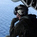 A military aircrew member is seated on the open ramp or door of an aircraft in flight over open ocean. The individual is wearing full flight gear including a helmet with visor, flight suit, and communications equipment, and appears to be scanning or observing the water below. The blue ocean is clearly visible in the background, suggesting the aircraft is at a moderate altitude.