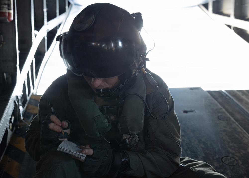 A military aircrew member wearing a flight helmet with visor and communications headset is seated inside what appears to be a military transport aircraft, writing notes on a small notepad. The individual is dressed in an olive drab flight suit with harness straps visible. The open ramp or door of the aircraft is visible in the background, with bright light suggesting flight conditions.