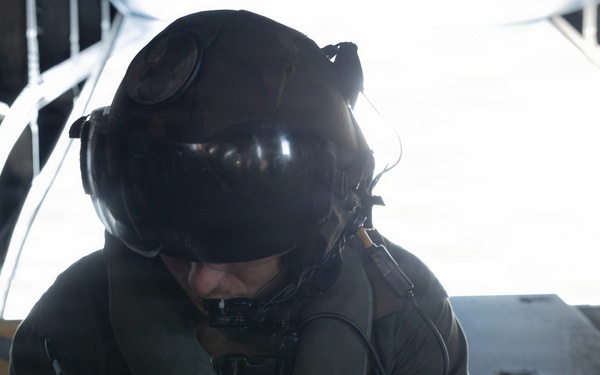 A military aircrew member wearing a flight helmet with visor and communications headset is seated inside what appears to be a military transport aircraft, writing notes on a small notepad. The individual is dressed in an olive drab flight suit with harness straps visible. The open ramp or door of the aircraft is visible in the background, with bright light suggesting flight conditions.