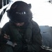 A military aircrew member wearing a flight helmet with visor and communications headset is seated inside what appears to be a military transport aircraft, writing notes on a small notepad. The individual is dressed in an olive drab flight suit with harness straps visible. The open ramp or door of the aircraft is visible in the background, with bright light suggesting flight conditions.