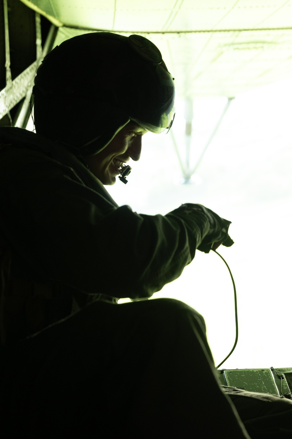 A silhouetted figure is seated inside what appears to be a military aircraft, backlit by bright light coming through an open cargo door or ramp. The person is wearing a helmet with night-vision goggle mounts and appears to be holding a cable or cord. The strong contrast between the dark interior and bright exterior creates a dramatic, high-contrast composition.