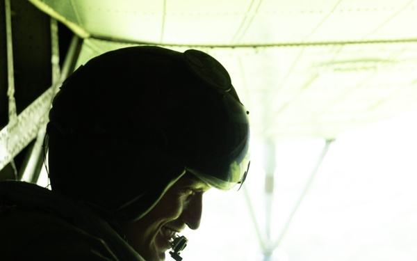 A silhouetted figure is seated inside what appears to be a military aircraft, backlit by bright light coming through an open cargo door or ramp. The person is wearing a helmet with night-vision goggle mounts and appears to be holding a cable or cord. The strong contrast between the dark interior and bright exterior creates a dramatic, high-contrast composition.