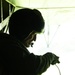 A silhouetted figure is seated inside what appears to be a military aircraft, backlit by bright light coming through an open cargo door or ramp. The person is wearing a helmet with night-vision goggle mounts and appears to be holding a cable or cord. The strong contrast between the dark interior and bright exterior creates a dramatic, high-contrast composition.