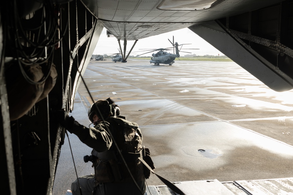 A military aircrew member in full flight gear and helmet is positioned at the open rear ramp of a large military helicopter or transport aircraft, looking out onto a wet tarmac. At least two large military helicopters, consistent with CH-53 Super Stallion variants, are visible on the flight line outside. The scene appears to depict pre-mission staging or flight operations at a military airfield.