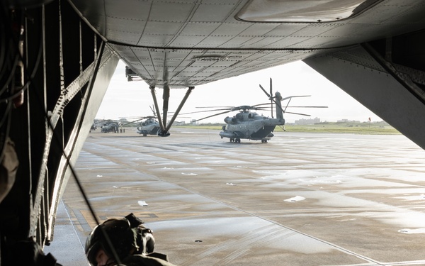 A military aircrew member in full flight gear and helmet is positioned at the open rear ramp of a large military helicopter or transport aircraft, looking out onto a wet tarmac. At least two large military helicopters, consistent with CH-53 Super Stallion variants, are visible on the flight line outside. The scene appears to depict pre-mission staging or flight operations at a military airfield.