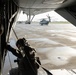 A military aircrew member in full flight gear and helmet is positioned at the open rear ramp of a large military helicopter or transport aircraft, looking out onto a wet tarmac. At least two large military helicopters, consistent with CH-53 Super Stallion variants, are visible on the flight line outside. The scene appears to depict pre-mission staging or flight operations at a military airfield.