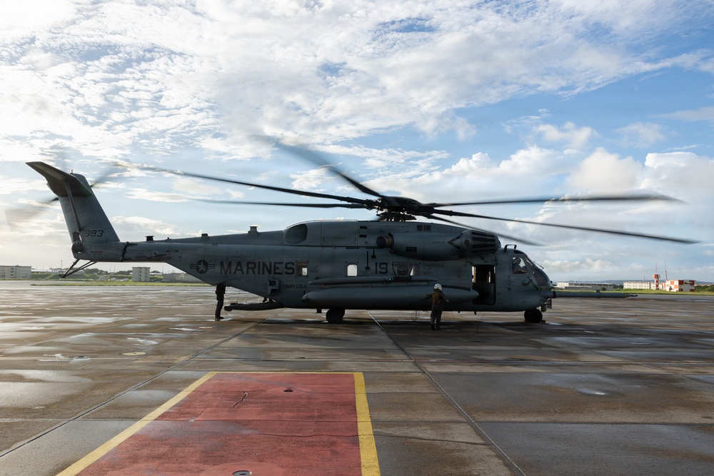 A U.S. Marines heavy-lift helicopter, marked 'MARINES' and bearing the number '19' and tail number '993', is parked on a wet tarmac with its rotors spinning. Two personnel are visible near the aircraft, one near the rear and one near the front cockpit area. The setting appears to be a military airfield, likely in a Pacific region based on the surrounding architecture.