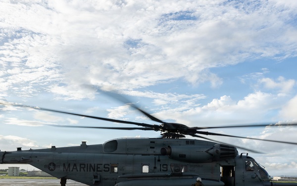 A U.S. Marines heavy-lift helicopter, marked 'MARINES' and bearing the number '19' and tail number '993', is parked on a wet tarmac with its rotors spinning. Two personnel are visible near the aircraft, one near the rear and one near the front cockpit area. The setting appears to be a military airfield, likely in a Pacific region based on the surrounding architecture.