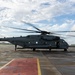 A U.S. Marines heavy-lift helicopter, marked 'MARINES' and bearing the number '19' and tail number '993', is parked on a wet tarmac with its rotors spinning. Two personnel are visible near the aircraft, one near the rear and one near the front cockpit area. The setting appears to be a military airfield, likely in a Pacific region based on the surrounding architecture.