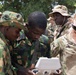 A group of military personnel in mixed camouflage uniforms are gathered outdoors examining what appears to be a drone controller or tablet device. The group includes soldiers wearing different camouflage patterns, suggesting a multinational military training or cooperation exercise. A soldier wearing sunglasses on the right appears to be instructing or demonstrating the device to the others.