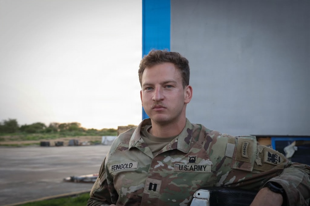 A U.S. Army soldier in Operational Camouflage Pattern (OCP) uniform is seated outdoors, looking directly at the camera with a neutral expression. His name tape reads 'FEINGOLD' and his chest tape reads 'U.S. ARMY.' He appears to be at a military installation or airfield, with open tarmac visible in the background.