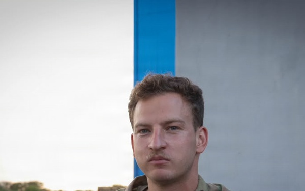 A U.S. Army soldier in Operational Camouflage Pattern (OCP) uniform is seated outdoors, looking directly at the camera with a neutral expression. His name tape reads 'FEINGOLD' and his chest tape reads 'U.S. ARMY.' He appears to be at a military installation or airfield, with open tarmac visible in the background.