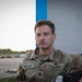 A U.S. Army soldier in Operational Camouflage Pattern (OCP) uniform is seated outdoors, looking directly at the camera with a neutral expression. His name tape reads 'FEINGOLD' and his chest tape reads 'U.S. ARMY.' He appears to be at a military installation or airfield, with open tarmac visible in the background.