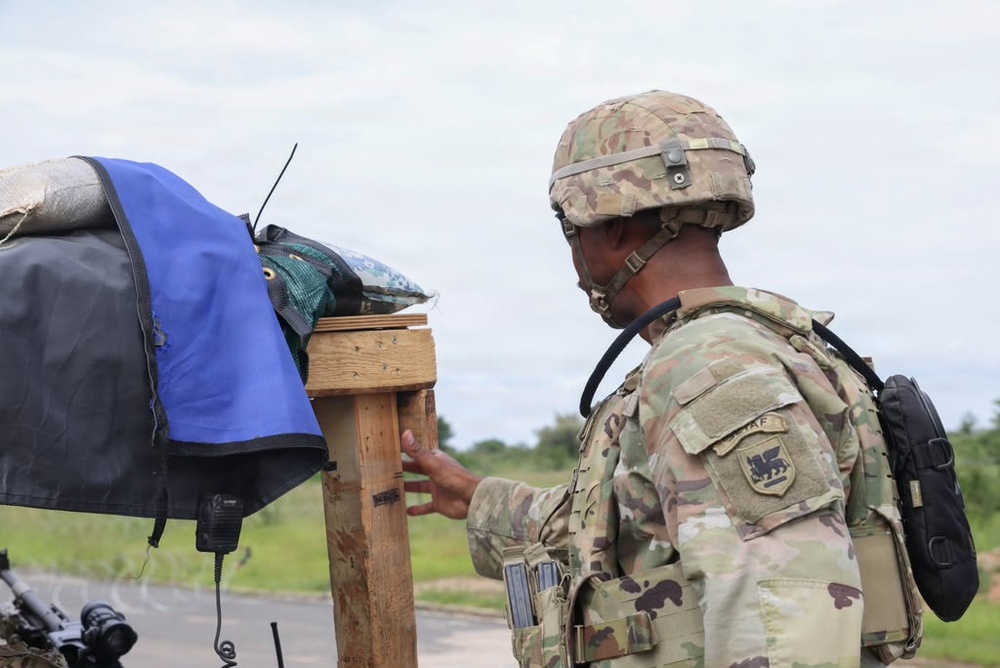 A soldier in full combat gear, wearing MultiCam/OCP camouflage uniform and helmet, is examining or interacting with a wooden post or stand that has various items draped over it including blue and black tarps or covers. The soldier has a unit patch visible on the left shoulder depicting what appears to be a griffin or dragon emblem. The setting appears to be an outdoor training or operational environment with a road and green vegetation visible in the background.
