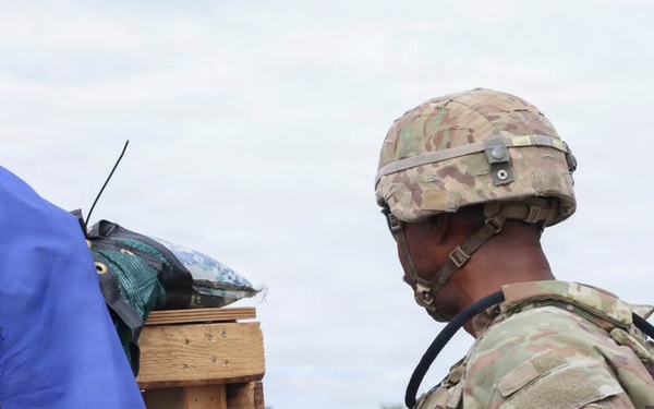 A soldier in full combat gear, wearing MultiCam/OCP camouflage uniform and helmet, is examining or interacting with a wooden post or stand that has various items draped over it including blue and black tarps or covers. The soldier has a unit patch visible on the left shoulder depicting what appears to be a griffin or dragon emblem. The setting appears to be an outdoor training or operational environment with a road and green vegetation visible in the background.
