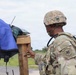 A soldier in full combat gear, wearing MultiCam/OCP camouflage uniform and helmet, is examining or interacting with a wooden post or stand that has various items draped over it including blue and black tarps or covers. The soldier has a unit patch visible on the left shoulder depicting what appears to be a griffin or dragon emblem. The setting appears to be an outdoor training or operational environment with a road and green vegetation visible in the background.
