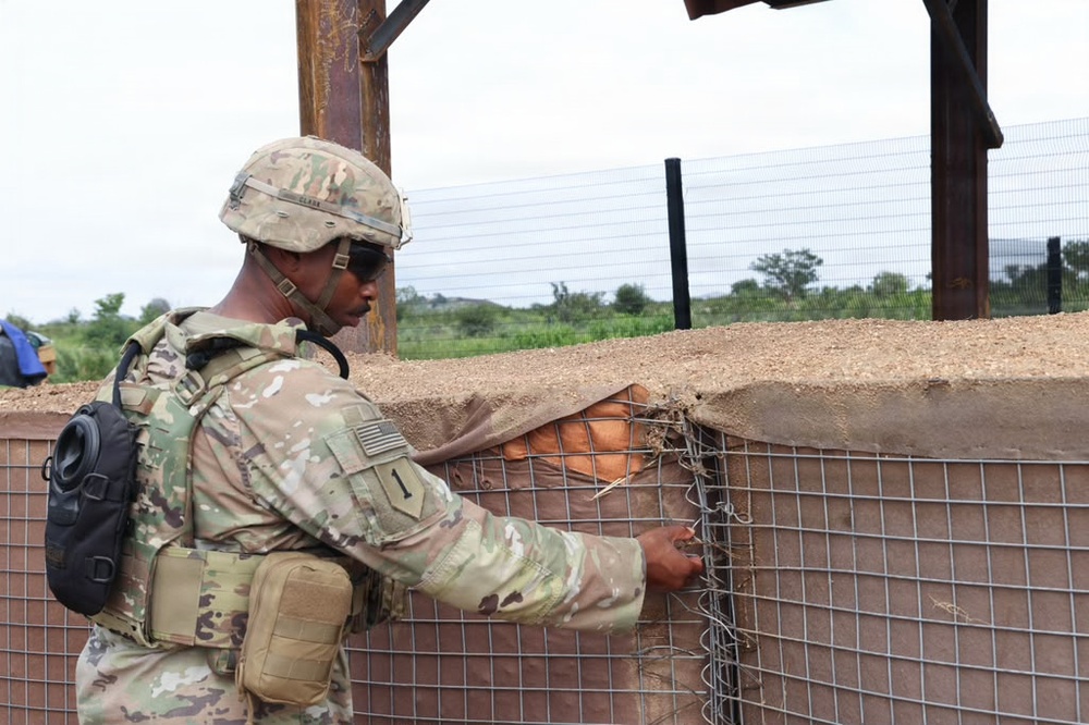 A U.S. Army soldier in full combat gear is inspecting or working on a wire mesh barrier attached to a fortified wall structure. The soldier is wearing Operational Camouflage Pattern (OCP) uniform with a helmet, body armor, and tactical accessories. The setting appears to be an outdoor military installation or forward operating base.