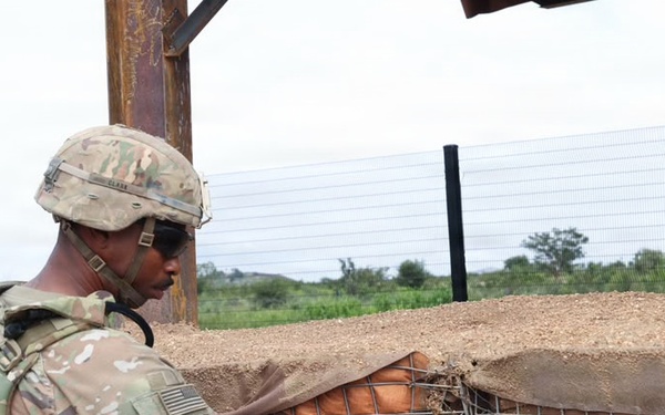 A U.S. Army soldier in full combat gear is inspecting or working on a wire mesh barrier attached to a fortified wall structure. The soldier is wearing Operational Camouflage Pattern (OCP) uniform with a helmet, body armor, and tactical accessories. The setting appears to be an outdoor military installation or forward operating base.