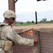 A U.S. Army soldier in full combat gear is inspecting or working on a wire mesh barrier attached to a fortified wall structure. The soldier is wearing Operational Camouflage Pattern (OCP) uniform with a helmet, body armor, and tactical accessories. The setting appears to be an outdoor military installation or forward operating base.