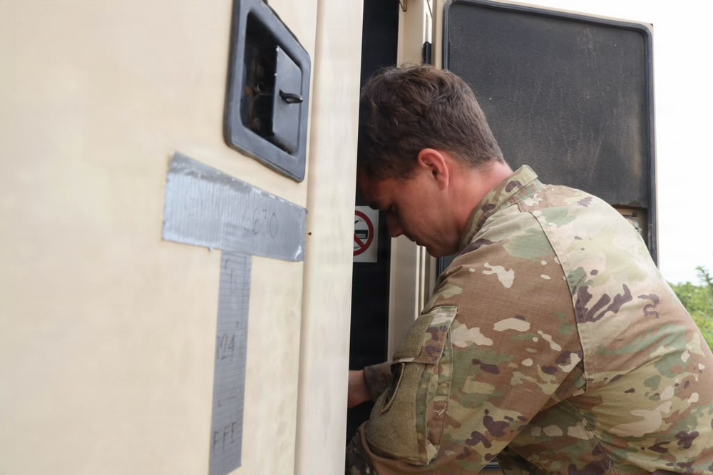 A military service member in MultiCam/OCP camouflage uniform is leaning into or inspecting the doorway of a military vehicle or shelter unit. The vehicle exterior is light tan/beige and has duct tape markings with handwritten text visible on the side. The scene appears to be an outdoor military operational environment.