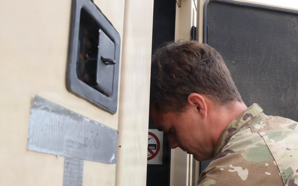 A military service member in MultiCam/OCP camouflage uniform is leaning into or inspecting the doorway of a military vehicle or shelter unit. The vehicle exterior is light tan/beige and has duct tape markings with handwritten text visible on the side. The scene appears to be an outdoor military operational environment.