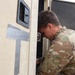 A military service member in MultiCam/OCP camouflage uniform is leaning into or inspecting the doorway of a military vehicle or shelter unit. The vehicle exterior is light tan/beige and has duct tape markings with handwritten text visible on the side. The scene appears to be an outdoor military operational environment.