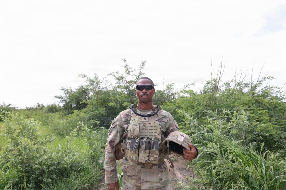 A U.S. Army soldier in full MultiCam/OCP camouflage uniform and plate carrier vest stands in a lush, green outdoor environment. He is holding a combat helmet in his right hand and wearing tactical sunglasses. The setting appears to be a field or rural area with dense vegetation in the background.