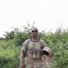 A U.S. Army soldier in full MultiCam/OCP camouflage uniform and plate carrier vest stands in a lush, green outdoor environment. He is holding a combat helmet in his right hand and wearing tactical sunglasses. The setting appears to be a field or rural area with dense vegetation in the background.