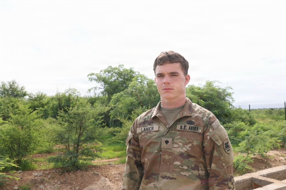 A young U.S. Army soldier wearing OCP (Operational Camouflage Pattern) uniform is photographed outdoors. His name tape reads 'LANIER' and the branch tape reads 'U.S. ARMY'. The background shows green shrubs/trees and an overcast sky, suggesting a semi-arid or tropical environment.