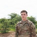 A young U.S. Army soldier wearing OCP (Operational Camouflage Pattern) uniform is photographed outdoors. His name tape reads 'LANIER' and the branch tape reads 'U.S. ARMY'. The background shows green shrubs/trees and an overcast sky, suggesting a semi-arid or tropical environment.