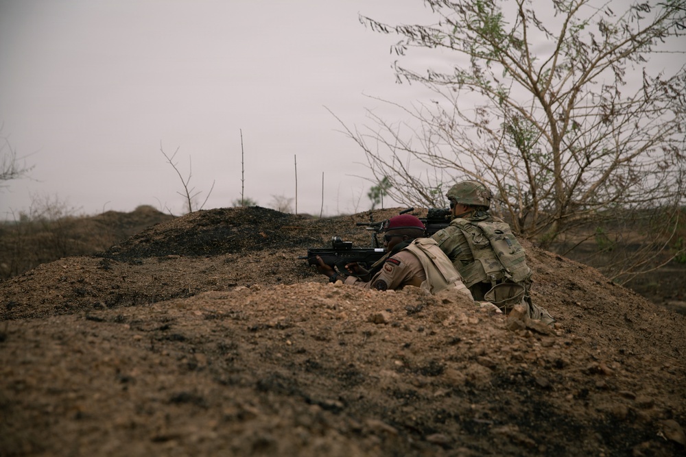 Two soldiers are positioned in a prone/crouching stance behind an earthen berm, aiming weapons downrange in an arid, semi-arid environment. One soldier wears a maroon beret and tan/khaki uniform, while the other wears a camouflage uniform with a helmet and tactical backpack. The setting appears to be a training exercise or operational position in a dry, sparse landscape.