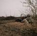 Two soldiers are positioned in a prone/crouching stance behind an earthen berm, aiming weapons downrange in an arid, semi-arid environment. One soldier wears a maroon beret and tan/khaki uniform, while the other wears a camouflage uniform with a helmet and tactical backpack. The setting appears to be a training exercise or operational position in a dry, sparse landscape.