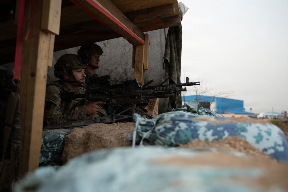 Two soldiers in military combat gear are positioned behind a fortified sandbag emplacement, with one aiming a machine gun outward through an opening. They are sheltered under a wooden-framed structure with tarp covering, suggesting a defensive fighting position. The setting appears to be a forward operating base or training exercise environment.