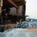 Two soldiers in military combat gear are positioned behind a fortified sandbag emplacement, with one aiming a machine gun outward through an opening. They are sheltered under a wooden-framed structure with tarp covering, suggesting a defensive fighting position. The setting appears to be a forward operating base or training exercise environment.