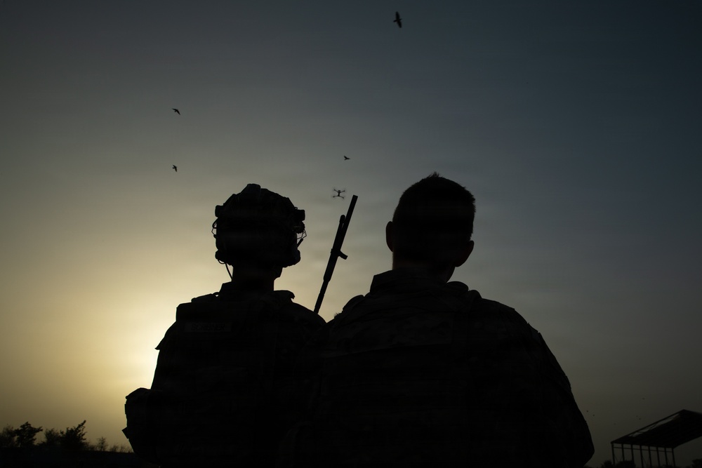 A silhouetted image of two individuals photographed from behind against a sunset sky. One figure is wearing a military-style helmet and tactical gear with what appears to be a rifle or long weapon visible. Several birds are visible in flight across the sky.