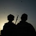 A silhouetted image of two individuals photographed from behind against a sunset sky. One figure is wearing a military-style helmet and tactical gear with what appears to be a rifle or long weapon visible. Several birds are visible in flight across the sky.