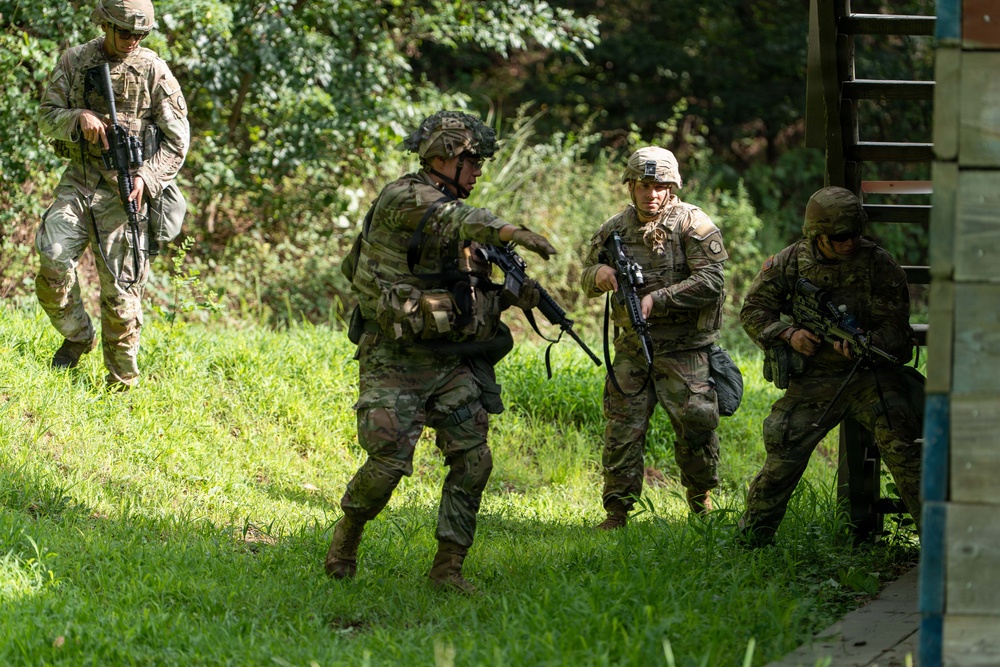 Four soldiers in camouflage uniforms and tactical gear are conducting what appears to be a military training exercise in a vegetated outdoor environment. The soldiers are armed with rifles and appear to be practicing building approach or clearing techniques near a structure. One soldier appears to be directing or signaling others, suggesting a coordinated tactical movement drill.