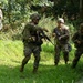Four soldiers in camouflage uniforms and tactical gear are conducting what appears to be a military training exercise in a vegetated outdoor environment. The soldiers are armed with rifles and appear to be practicing building approach or clearing techniques near a structure. One soldier appears to be directing or signaling others, suggesting a coordinated tactical movement drill.