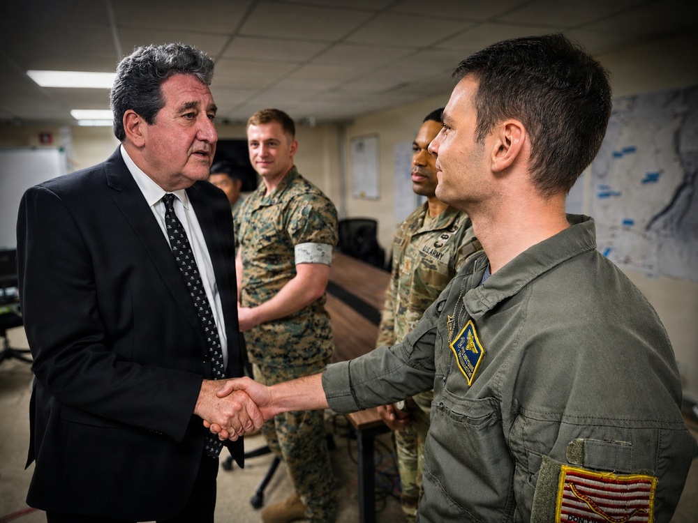 A civilian in a dark suit is shaking hands with a military officer wearing an olive green flight suit bearing patches including a U.S. flag. In the background, several other military personnel in various uniforms, including U.S. Marine Corps MARPAT camouflage and U.S. Army OCP uniforms, are observing the interaction. The setting appears to be an indoor military facility or briefing room.