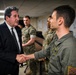 A civilian in a dark suit is shaking hands with a military officer wearing an olive green flight suit bearing patches including a U.S. flag. In the background, several other military personnel in various uniforms, including U.S. Marine Corps MARPAT camouflage and U.S. Army OCP uniforms, are observing the interaction. The setting appears to be an indoor military facility or briefing room.