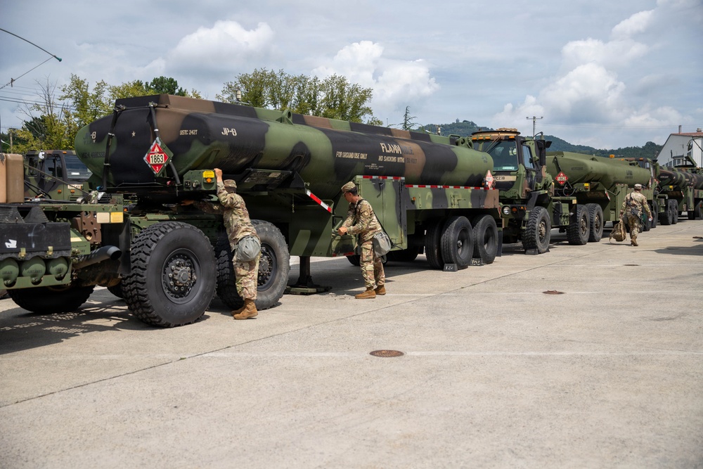 Military personnel in camouflage uniforms are working alongside a convoy of large military fuel tanker trucks in a staging area. The tankers are marked with hazardous materials placards (UN 1863) indicating jet fuel (JP-8), and bear 'FLAMMABLE' warnings. The scene appears to be a pre-mission preparation or equipment inspection at a military installation.