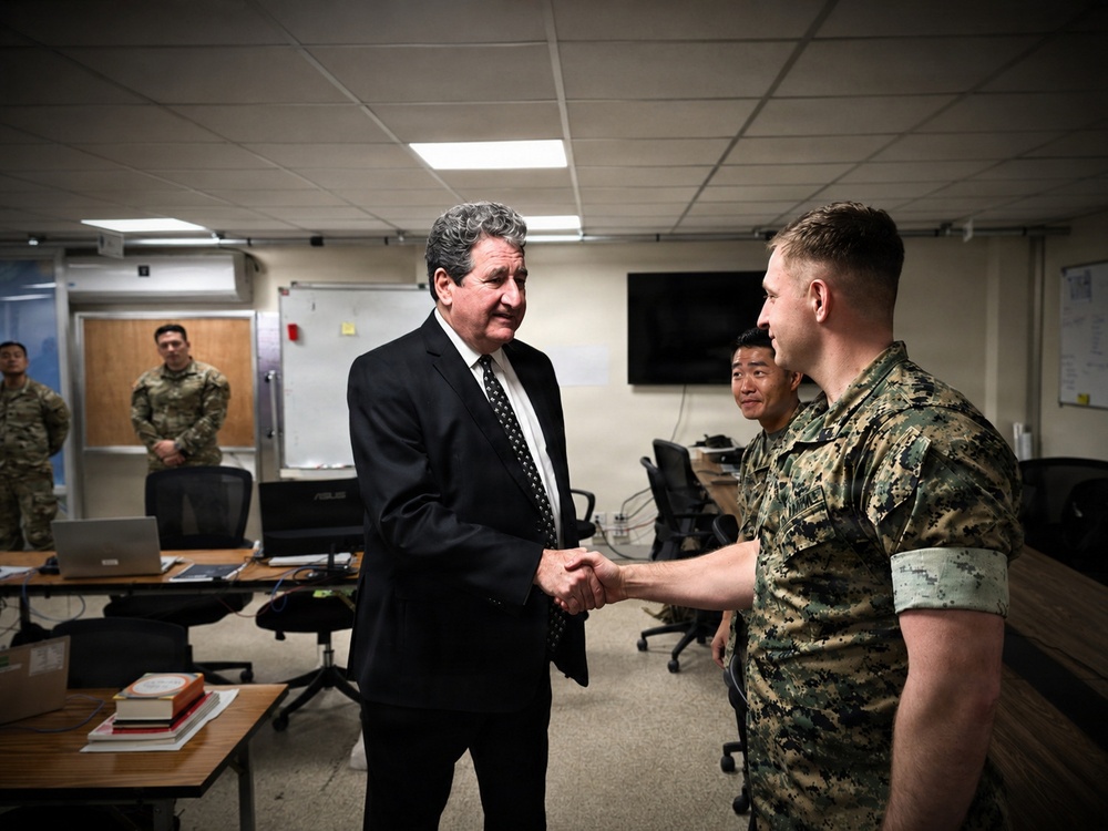 A civilian in a dark suit is shaking hands with a military personnel wearing Marine Corps digital camouflage (MARPAT) uniform in what appears to be a military operations or briefing room. A second military personnel in similar camouflage stands nearby observing, while two additional uniformed soldiers stand in the background near a doorway. The setting suggests an official visit or meeting at a military installation.