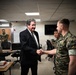 A civilian in a dark suit is shaking hands with a military personnel wearing Marine Corps digital camouflage (MARPAT) uniform in what appears to be a military operations or briefing room. A second military personnel in similar camouflage stands nearby observing, while two additional uniformed soldiers stand in the background near a doorway. The setting suggests an official visit or meeting at a military installation.