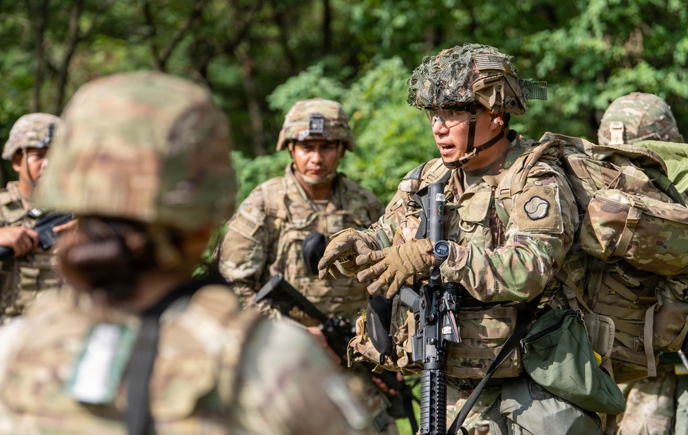 A group of military personnel in full combat gear are gathered in an outdoor wooded area, appearing to receive a briefing or instructions from a central soldier. The focal soldier is speaking and gesturing while holding an assault rifle, wearing a camouflage-netted helmet and full tactical kit. The scene appears to be a training exercise or operational briefing.