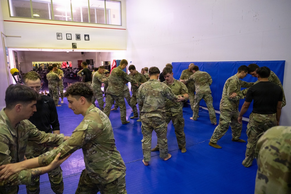 A group of military personnel in camouflage uniforms are conducting combatives or hand-to-hand combat training on blue gymnasium mats. The soldiers are paired up and practicing grappling or defensive techniques in what appears to be an indoor training facility. The setting includes wall-mounted crash pads and an adjacent weight room visible in the background.